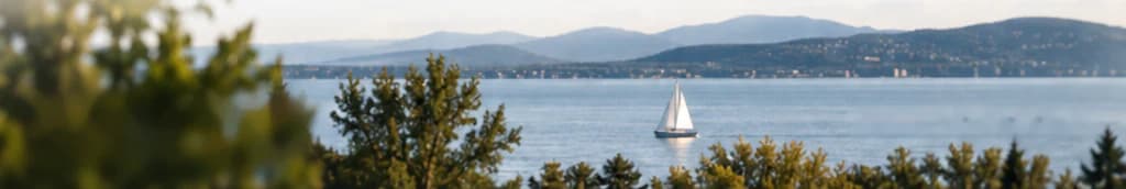 Panorama Danau Balaton dengan perahu layar, pohon-pohon pinus di latar depan, serta perbukitan dan permukiman di seberang pantai