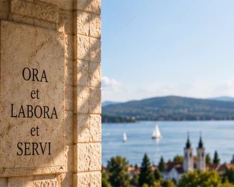Pilar batu bermandikan cahaya matahari dengan ukiran semboyan “ORA et LABORA et SERVI”, berlatarkan Danau Balaton berhias perahu layar, perbukitan, serta gereja bermenara dua