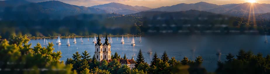 Cahaya senja keemasan di atas Danau Balaton: gereja biara putih bermenara dua berdiri di atas bukit berhutan, perahu layar di danau, jajaran bukit dan sinar matahari di latar belakang, pepohonan gelap di latar depan