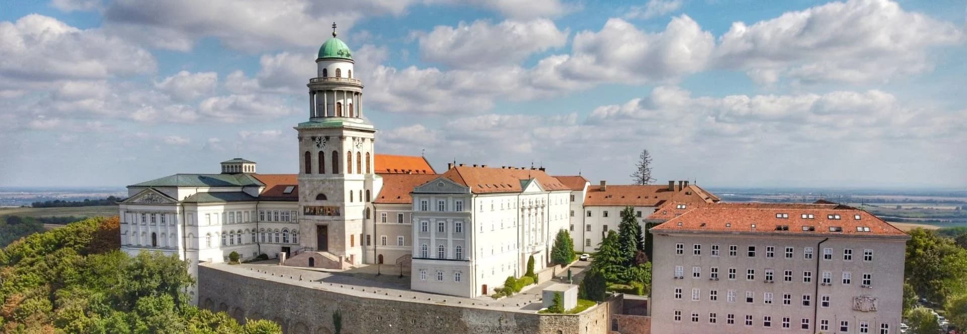 A Pannonhalmi Főapátság reggeli fényben: a zöld kupolás, keresztes torony és a vörös tetős, fehér épületszárnyak erdős dombtetőn, alatta a völgyben vörös tetős házak, a háttérben párás, ködös dombvonulatok.