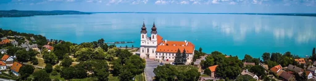 Alkonyi panoráma: balra erdős dombtetőn álló, tornyos fehér apátság, alatta lombos erdő, jobbra ciprusok és vörös tetős házak, a tavon két vitorlás, a túlparton kék dombvonulatok, a lemenő nap fénye a vízen csillog.