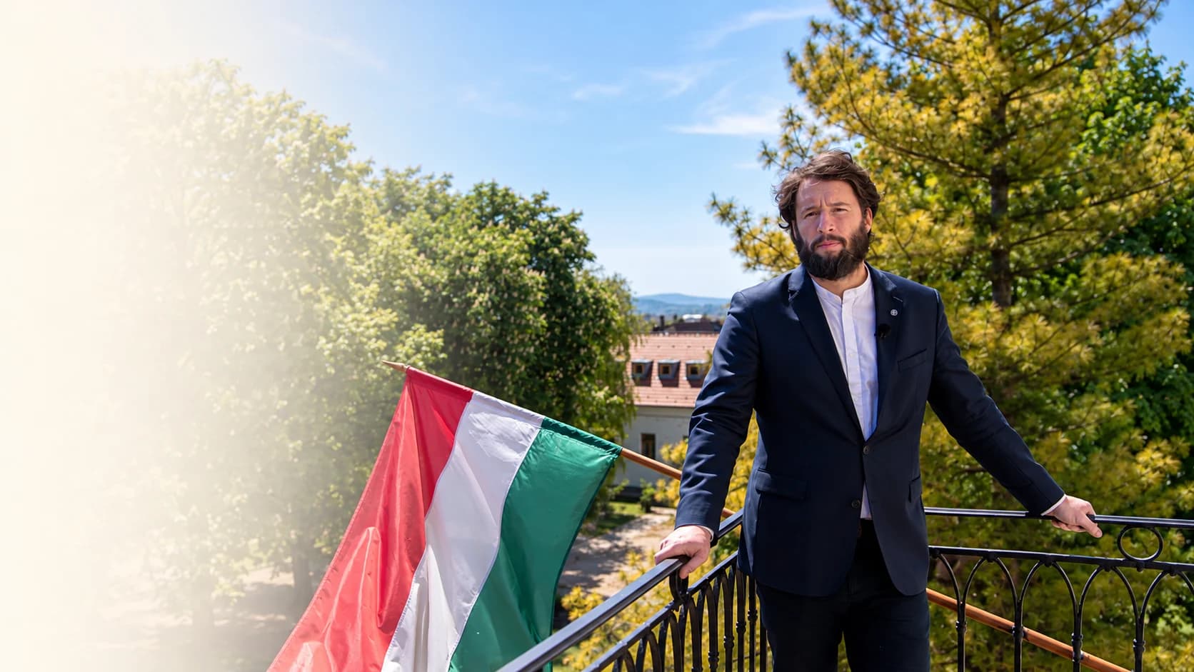 Kilátás egy napsütötte kőboltív alól a Balaton fölé magasodó kéttornyú bencés templomra, fenyők és lombos fák között, háttérben a tó és a kék dombok; jobbra kőpillér a vésett MÚLTBÓL ÉRTÉK – JELENBEN FELELŐSSÉG – JÖVŐNEK LEHETŐSÉG mottóval és egy kereszttel.