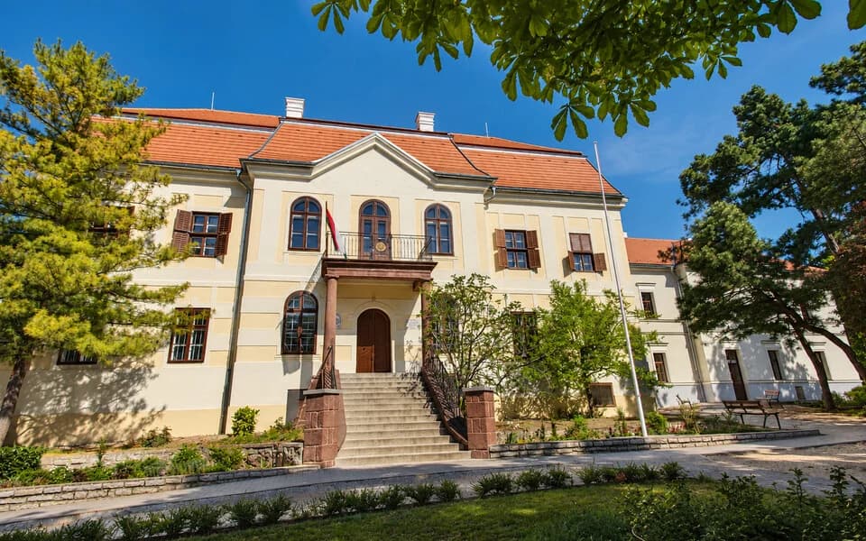 A krémszínű Kastély épület középső tornyával, előtte gyep és nagy fák