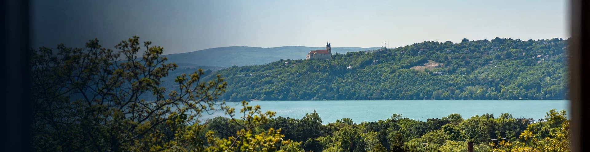 Kilátás a Balatonra vitorlásokkal és a túlparti dombokkal; balra elmosódott lombok, jobbra homokkő oszlopok az ORA et LABORA et SERVI vésett felirattal.
