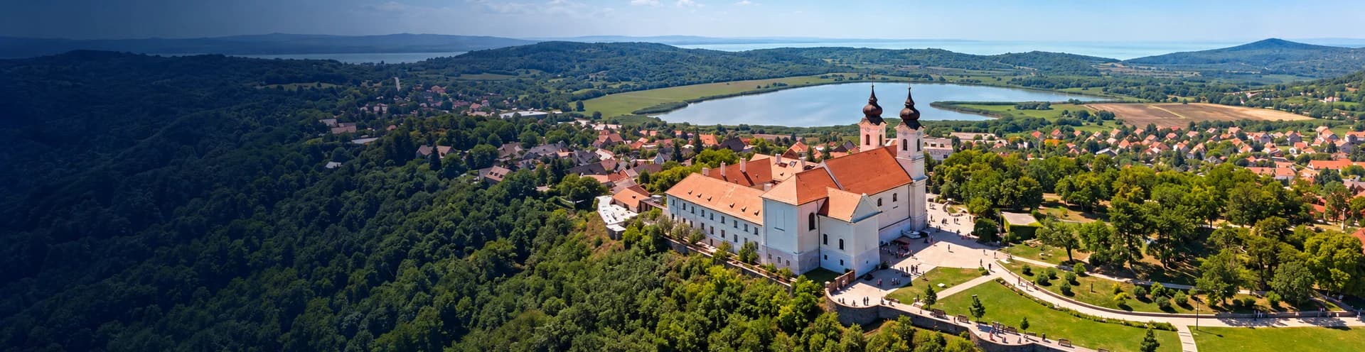 Naplementés panoráma a Balatonra vitorlásokkal, a túlparti dombokkal és egy kéttornyú templommal a fenyők között