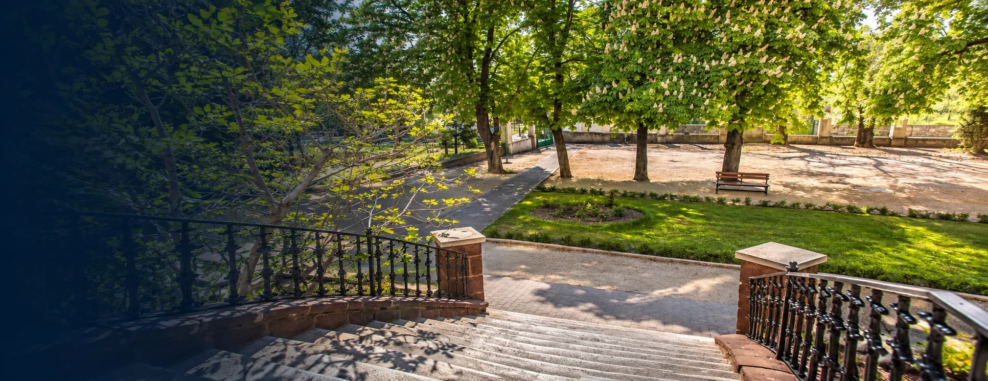 Panoráma egy erdős dombról a Balatonra: fenyők, piros tetők, kéttornyú templom, vitorlások a tavon, a túlparton kék hegyek