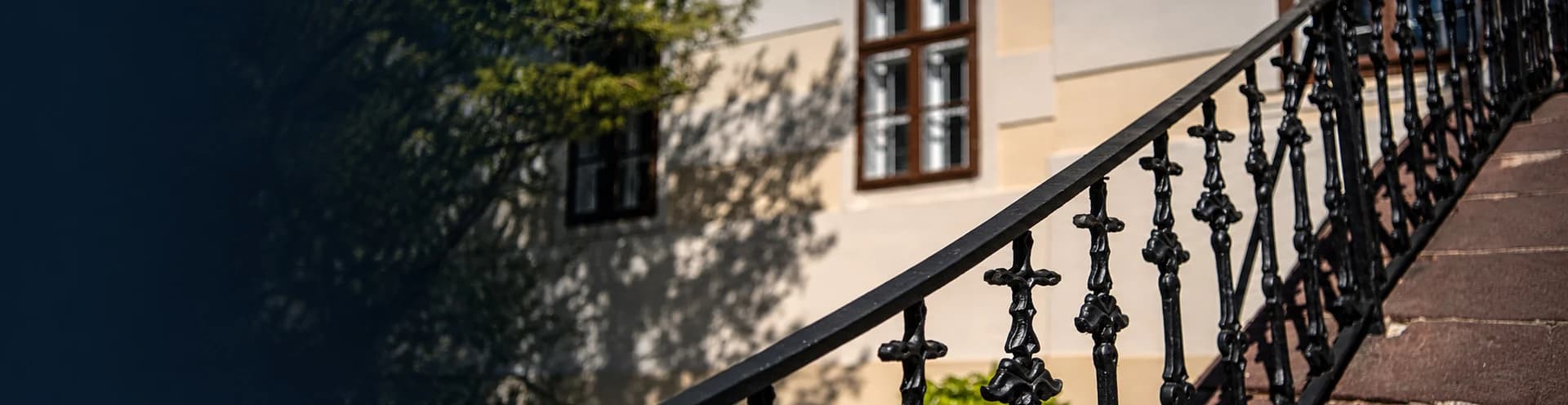 A Balaton délutáni fényben, vitorlásokkal a vízen, a túlparti dombokkal, előtérben napsütötte lombokkal és fenyőkkel