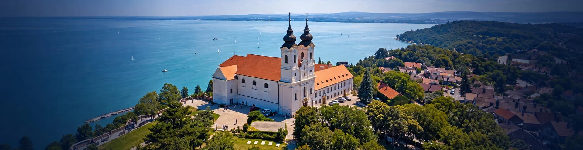 Aranyló esti fény a Balaton fölött: erdős dombon álló kéttornyú fehér apátsági templom, vitorlások a tavon, dombvonulatok és napsugarak a háttérben, sötét fák az előtérben